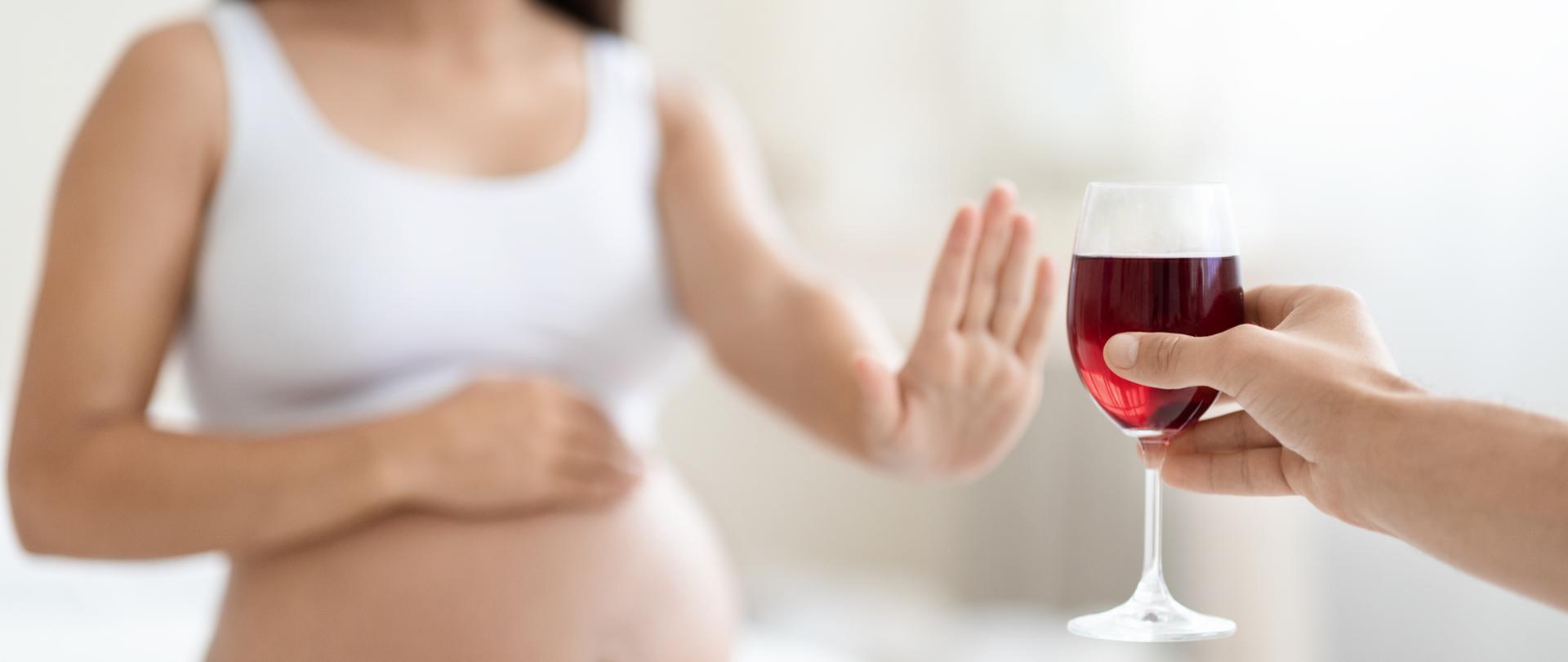 młoda kobieta w ciąży odmawia picia wina, wykonując gest zatrzymania w stronę szklanki, siedząc na łóżku w domu, dotykając brzucha