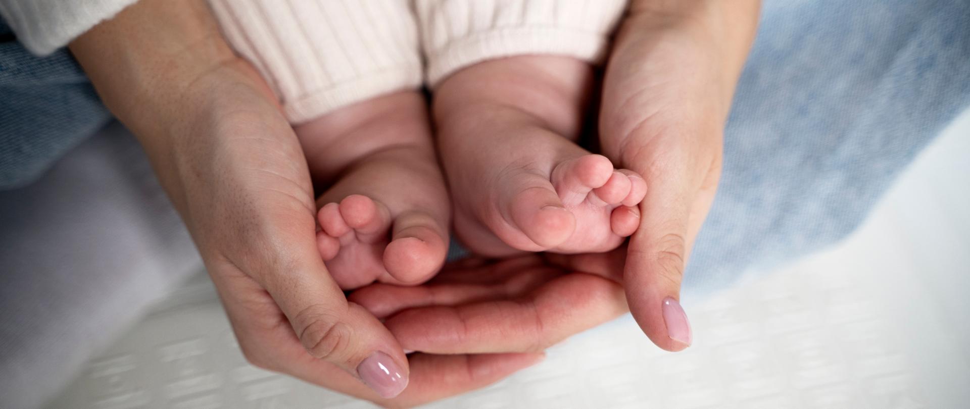 Zdjęcie przedstawia dłonie kobiety trzymające stopy małego dziecka