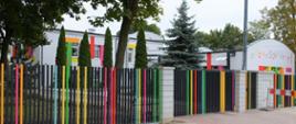 Na zdjęciu widać przedszkole nr 1 z ulicy przed nim czarny płot z kolorowymi częściami oraz różne drzewa.