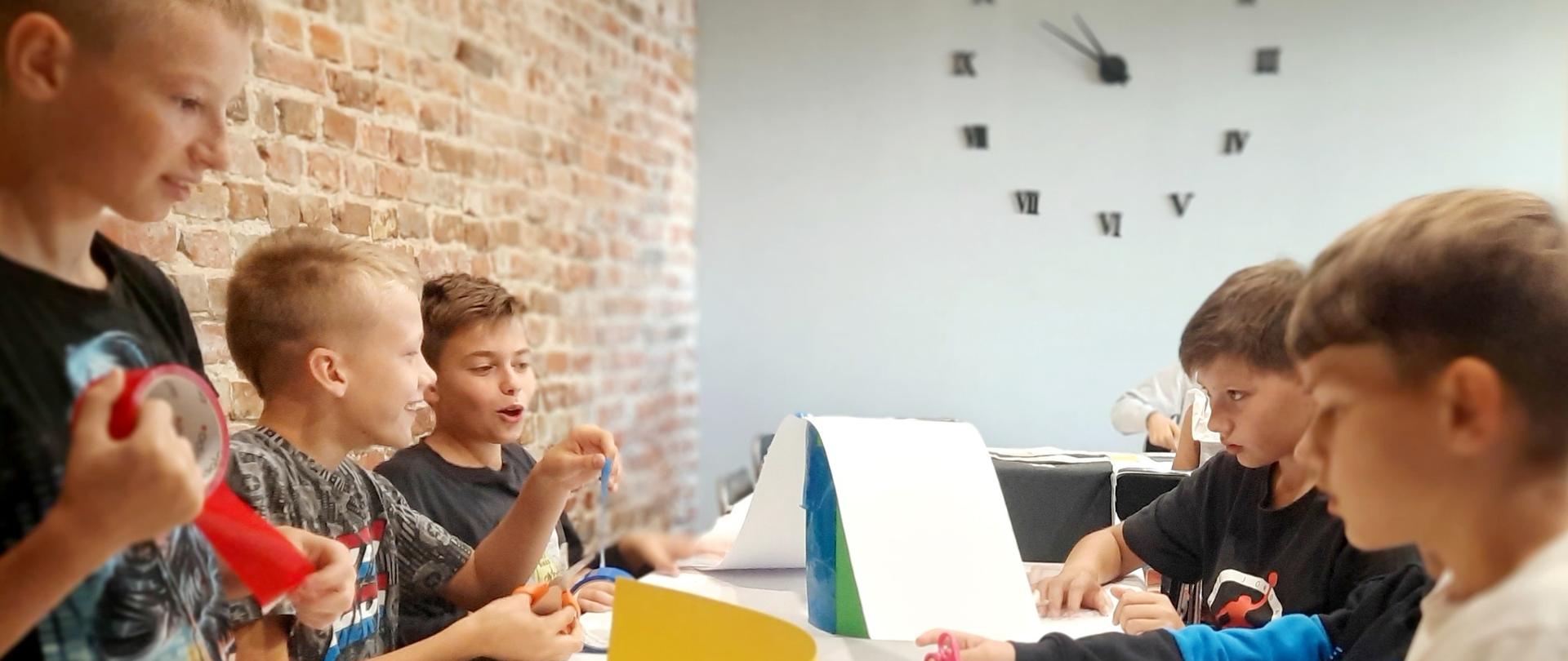 Pięciu chłopców w wieku około 10 lat przy stole w pracowni artystycznej tworzą pracę przestrzenną. W tle biała ściana, a na niej zegar.