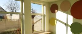 Sala w żłobku z zieloną podłogą na ścianie dookoła drzwi wiszą kolorowe koła. Po lewej stronie widać dwa okna.