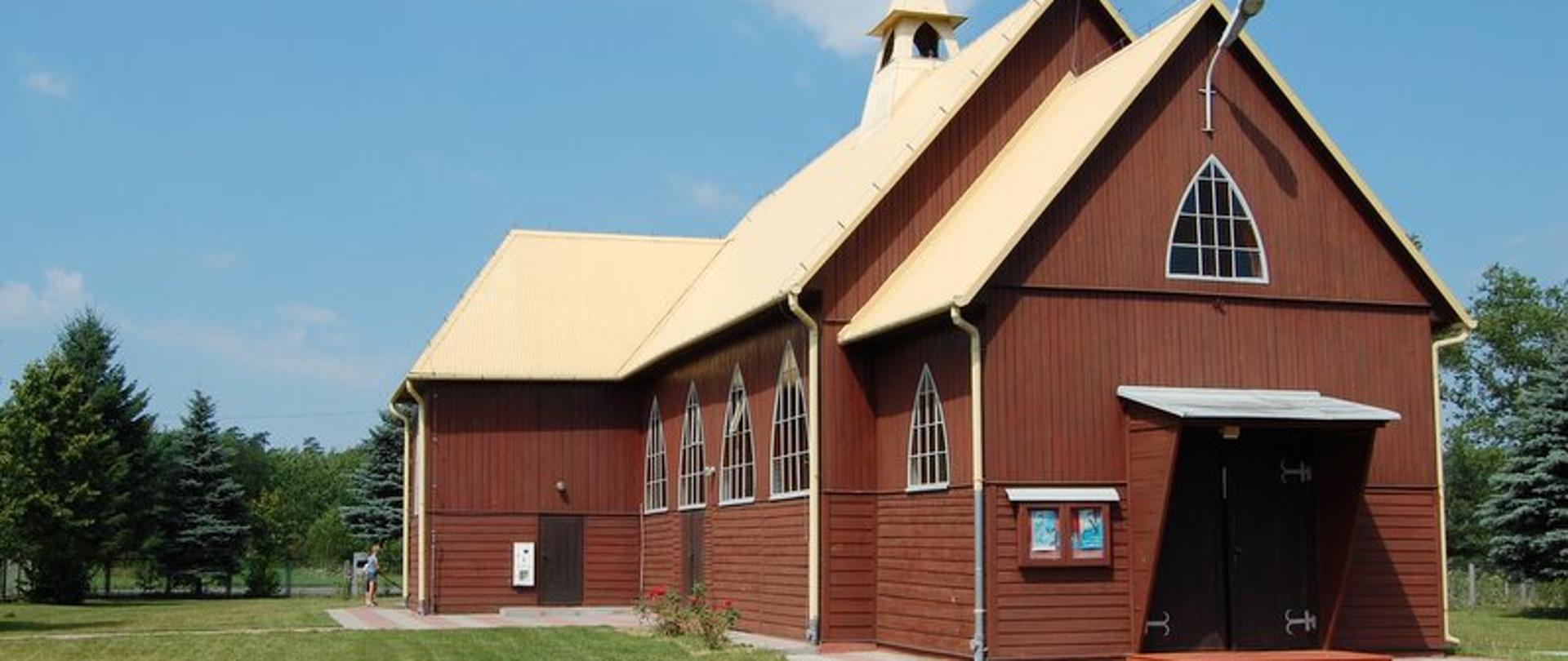 Drewniany, brązowy kościół z jasnym dachem, okna w kształcie trójkątów i duże brązowe drzwi. Przed kościołem zielony trawnik. 