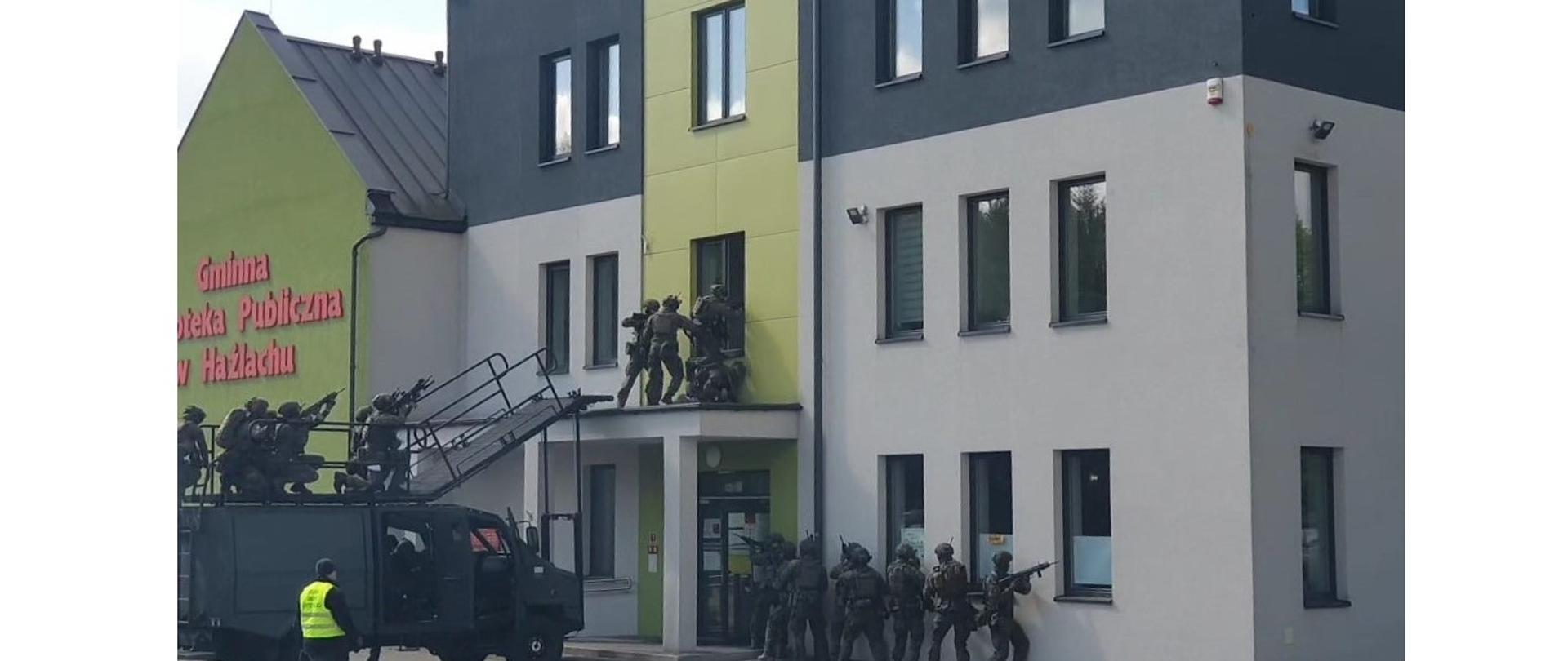 Wejście zespołu antyterrorystycznego na I piętro budynku GBP w Hażlachu. Przed budynkiem stoi specjalistyczny pojazd wojskowy z pomostem dla antyterrotystów. Uzbrojeni żołnierze wdzierają się do budynku przez okno. Druga grupa zajmuje pozycję przy ścianie budynku.
