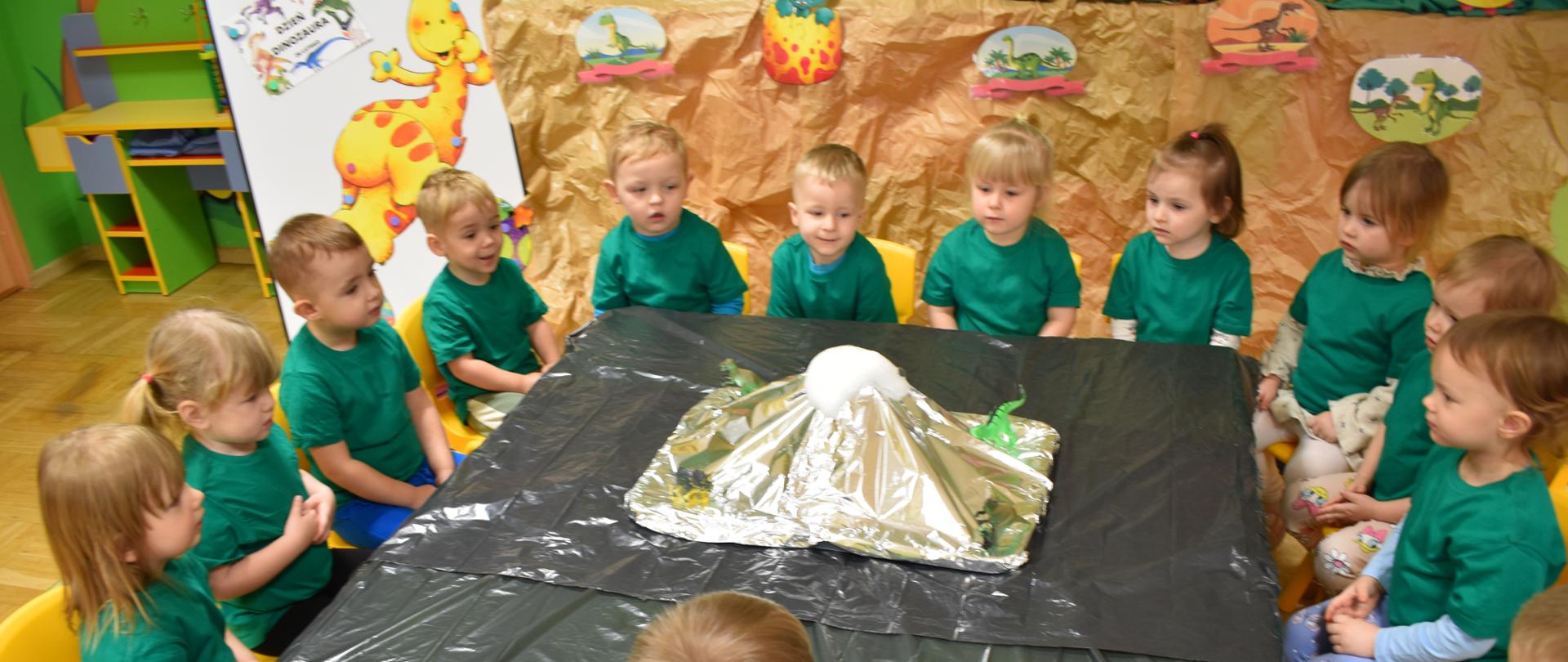 Na zdjęciu widać grupę dzieci siedzącą w koło stołu przykrytego czarną folią i obserwującą eksperyment wybuchającego wulkanu.