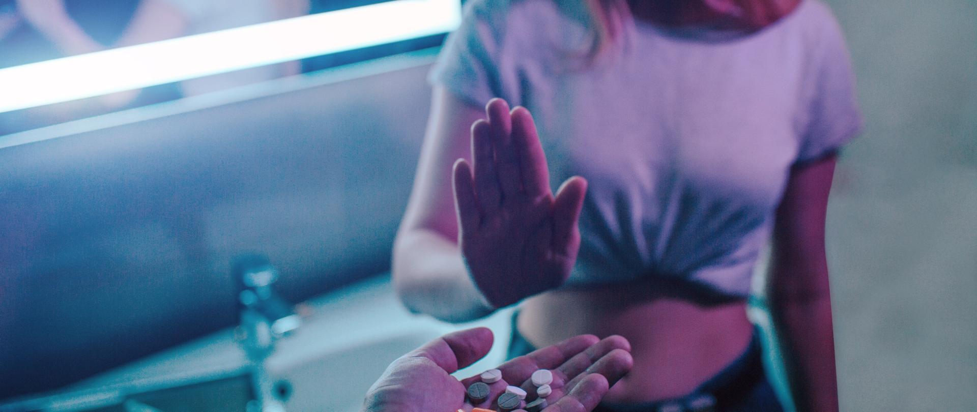 Żadnych narkotyków. Oferta leków. Odrzuć ofertę narkotyków. Ręka mówi NIE. Młoda kobieta pokazuje otwartą dłoń przeciwko ofercie narkotyków, trzymając w ręku kolorowe narkotyki w nocnym klubie. 