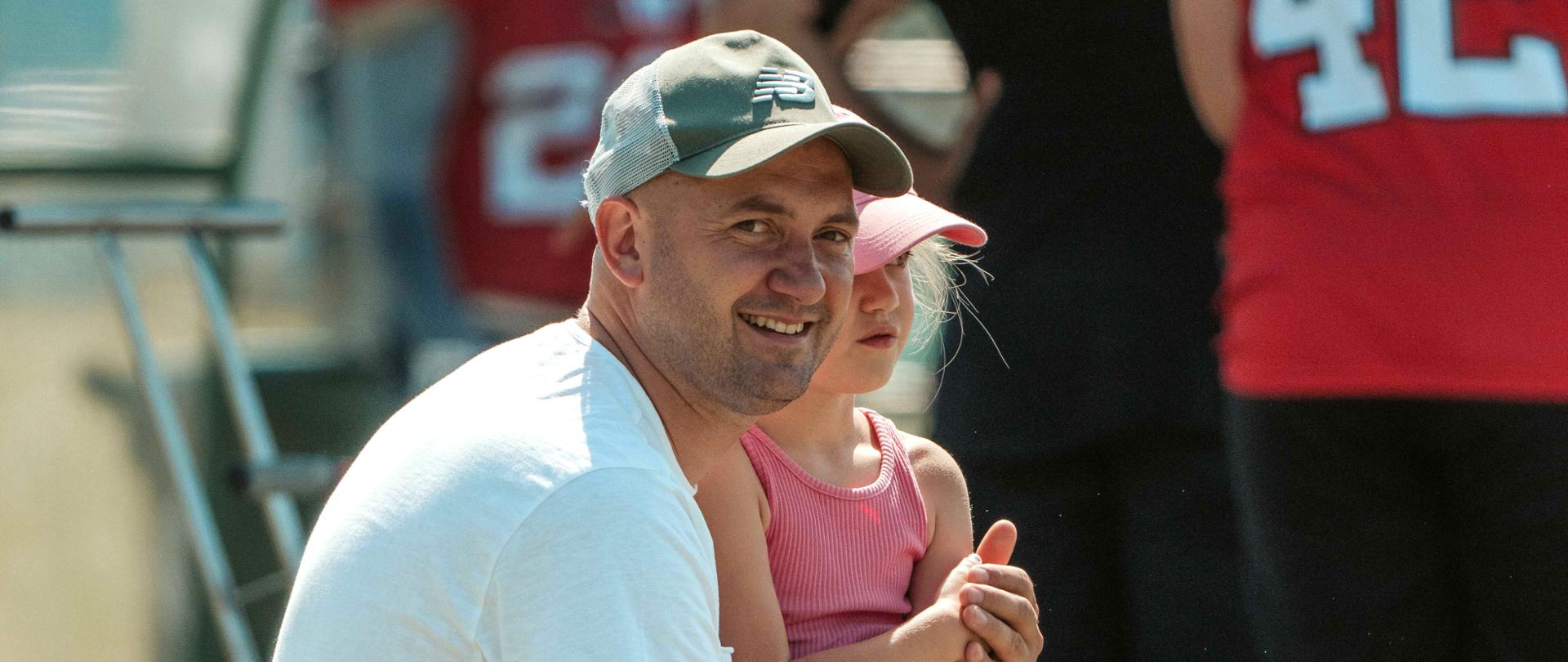 Ojciec z córką kucający na boisku, w tle zawodnicy w koszulkach z numerami.