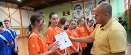 Grupka dziewczyn stoi na sali gimnastycznej, oraz mają na swoich szyjach medale, organizator w żółtej koszulce wręcza pierwszej dziewczynie dyplom gratulacyjny, uczęszczania w zawodach sportowych.