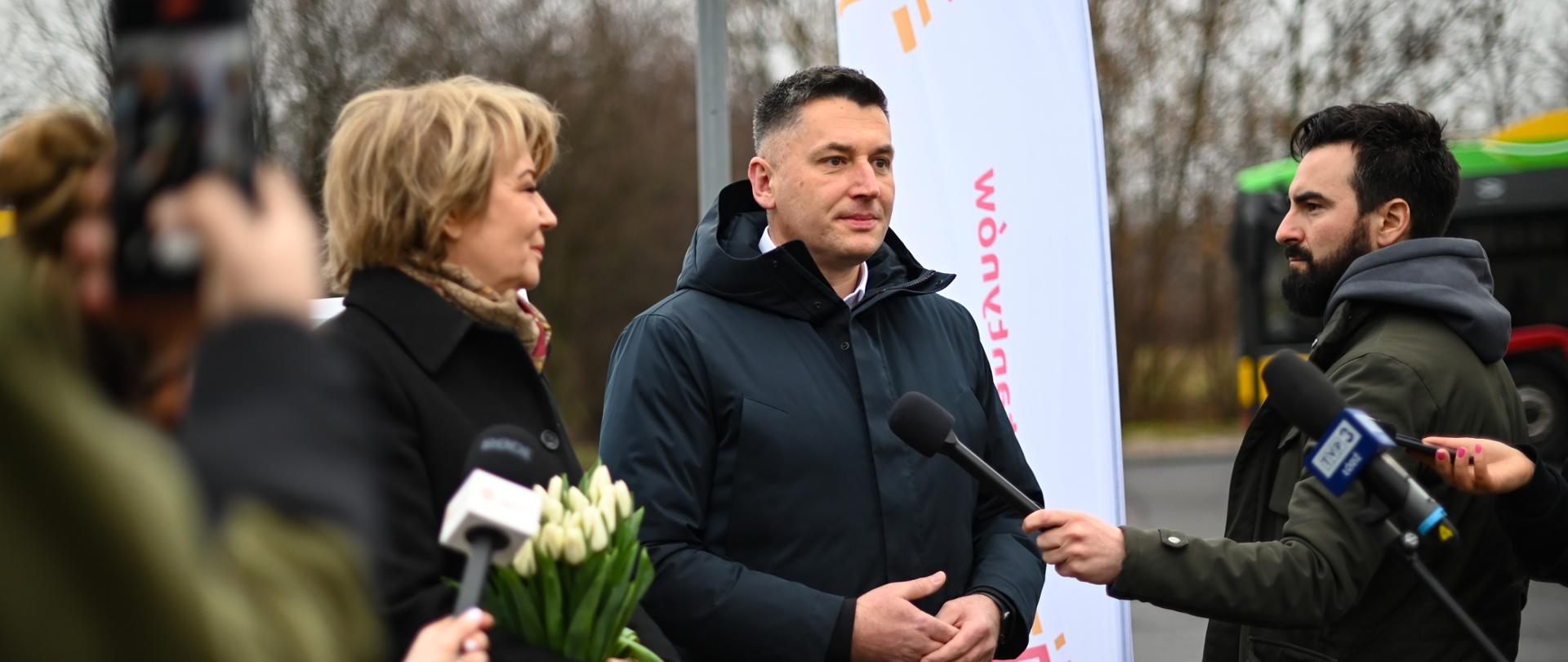 Zdjęcie z konferencji prasowej burmistrza Roberta Jakubowskiego i prezydent Hanny Zdanowskiej. W tle żagiel z logo Konstantynowa. Po bokach dziennikarze trzymający mikrofony.