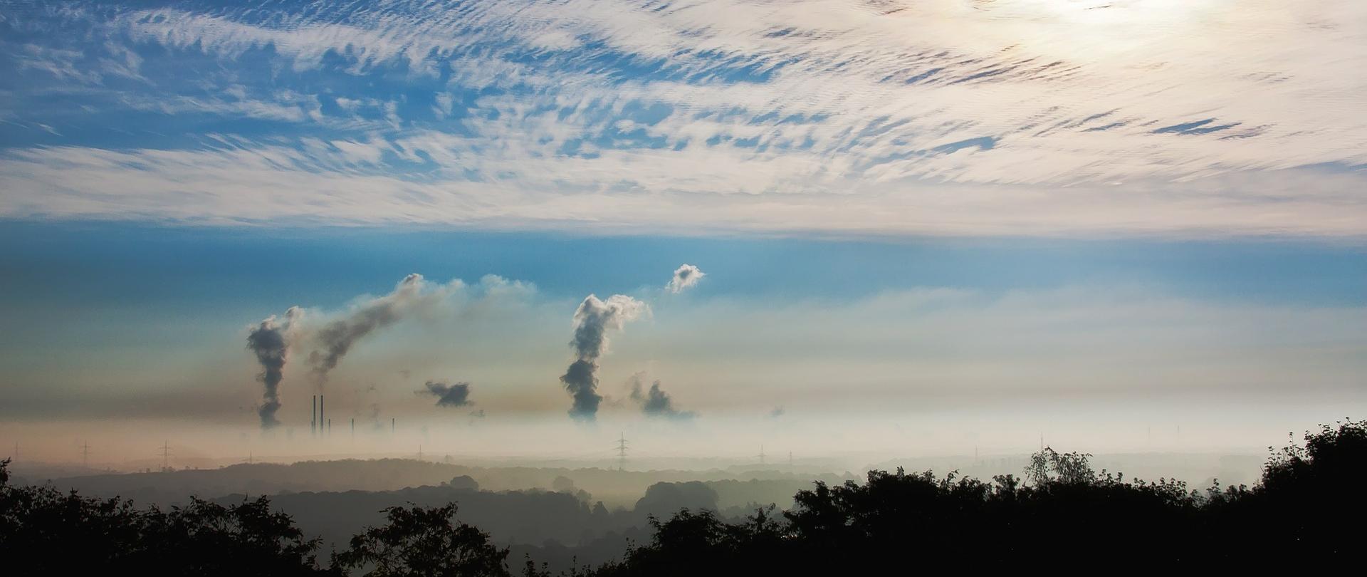 Widok na horyzont zasnuty dymem. Na pierwszym planie czubki drzew, w oddali kominy fabryk wystające spod warstwy smogu. Na niebie cienka warstwa chmur utrudniająca przedostanie się promieni słonecznych.