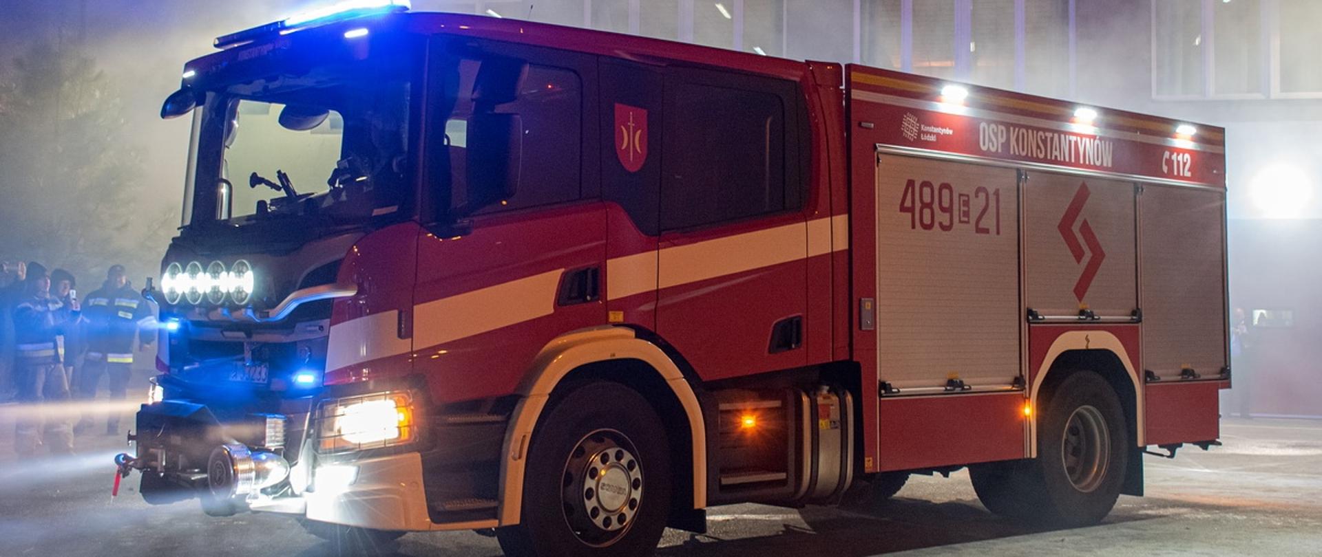 Zdjęcie zrobiono po zmroku. Przedstawia czerwony samochód strażacki z zapalonymi światłami. Wóz ratowniczo-gaśniczy. W tle budynek remizy ochotniczej straży pożarnej i grupka strażaków. Na samochodzie napis OSP Konstantynów, nr jednostki 489 21 i nr alarmowy 112