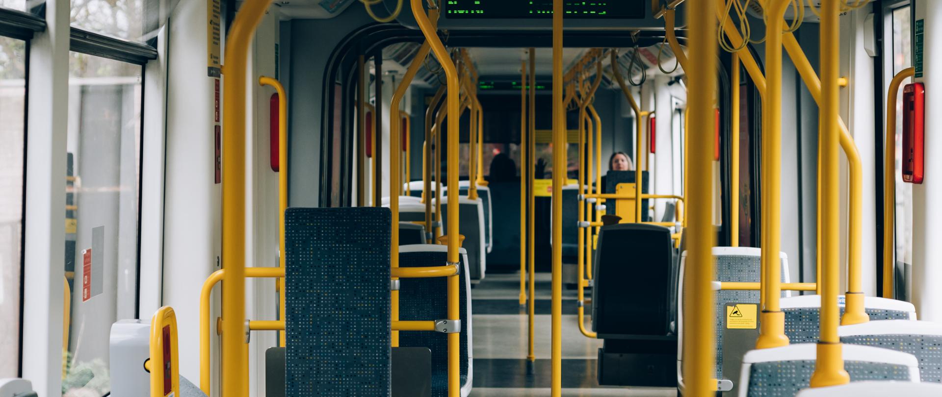 Wnętrze autobusu, szare fotele, żółte poręcze. 