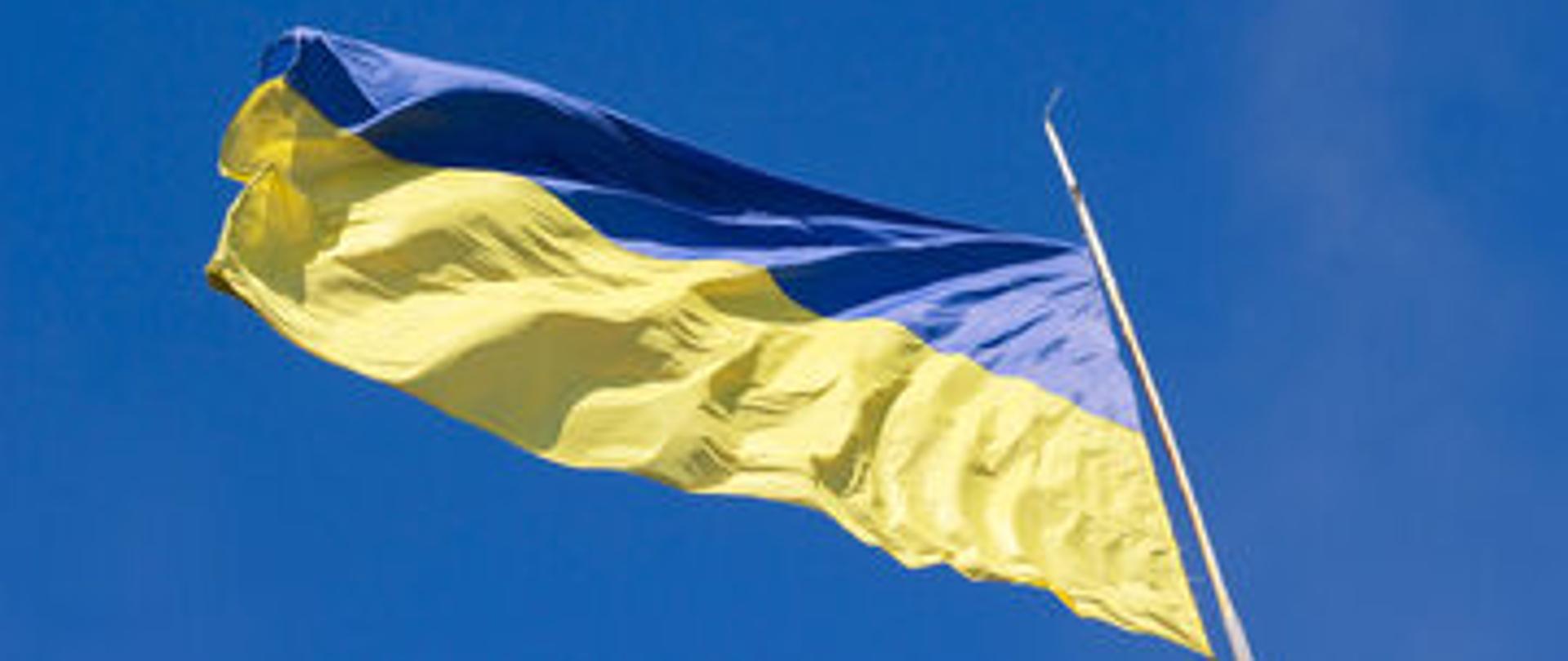 Niebiesko - żółta flaga Ukrainy powiewająca na białym maszcie na tle błękitnego nieba