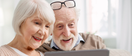 Zdjęcie przedstawia starszych, uśmiechniętych ludzi, którzy patrzą na laptopa. Kobieta mi siwe włosy oraz czerwone usta, a mężczyzna zarost i okulary na głowie.