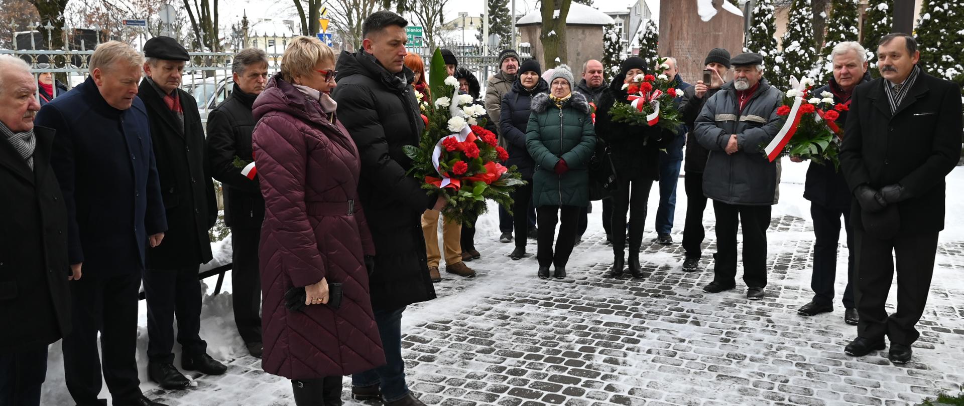 Burmistrz Konstantynowa Łódzkiego wraz z przewodniczącą Rady Miasta Jadwigą Czekajewską stoją obok siebie trzymając biało-czerwone kwiaty. Za nimi stoją ludzie, którzy przyglądają się im, trzymając również wiązanki.