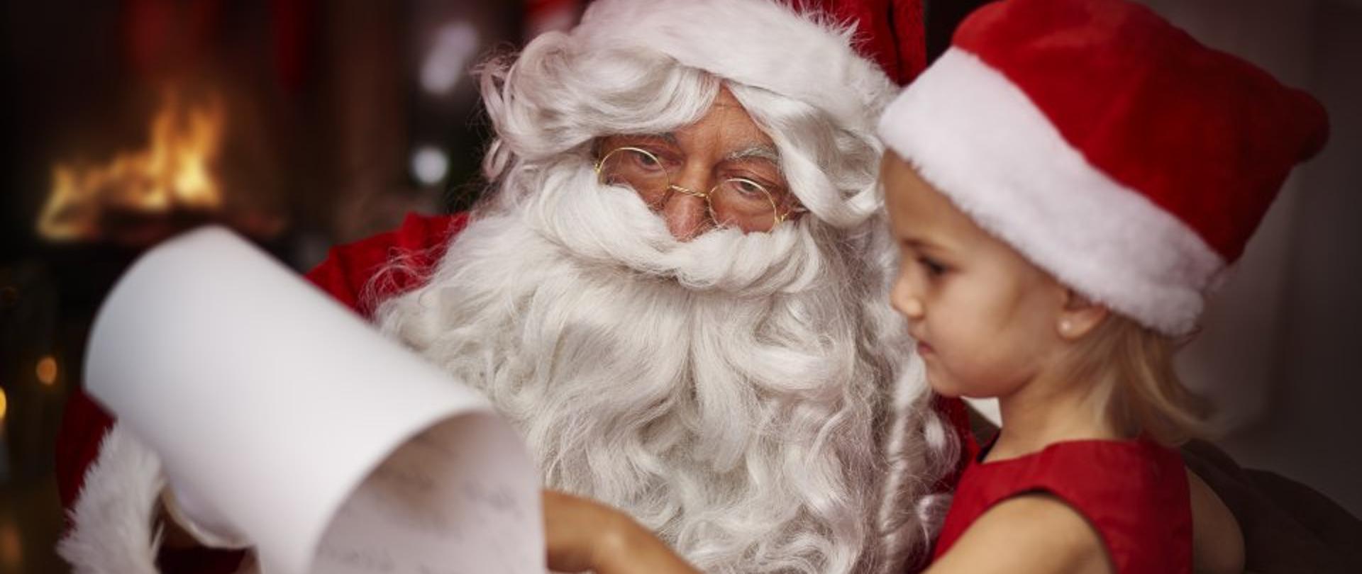 Zdjęcie przedstawia Świętego Mikołaja z długą listą prezentów. Obok niego siedzi ubrana w czapkę Mikołaja mała dziewczynka która wskazuje na jakąś pozycję na liście.