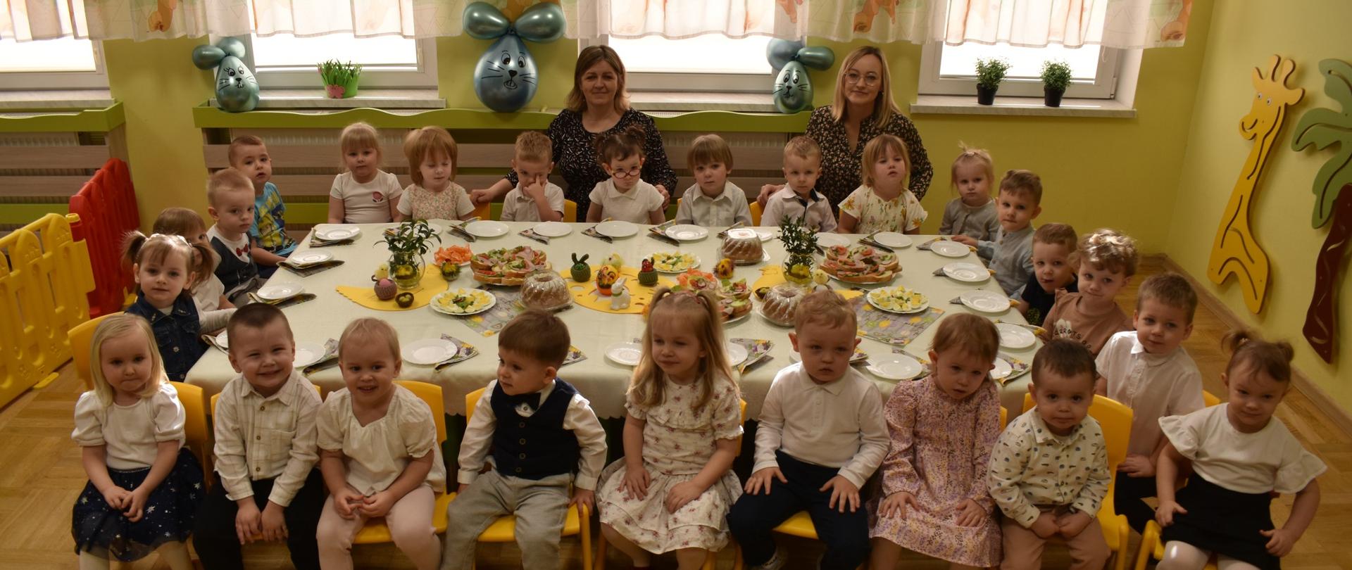 Na zdjęciu widać grupę dzieci siedzącą przy stole wielkanocnym. stół jest przykryty białym obrusem a na stole widać symbole i potrawy związane z Wielkanocą. za dziećmi siedzą trzy opoiekunki.