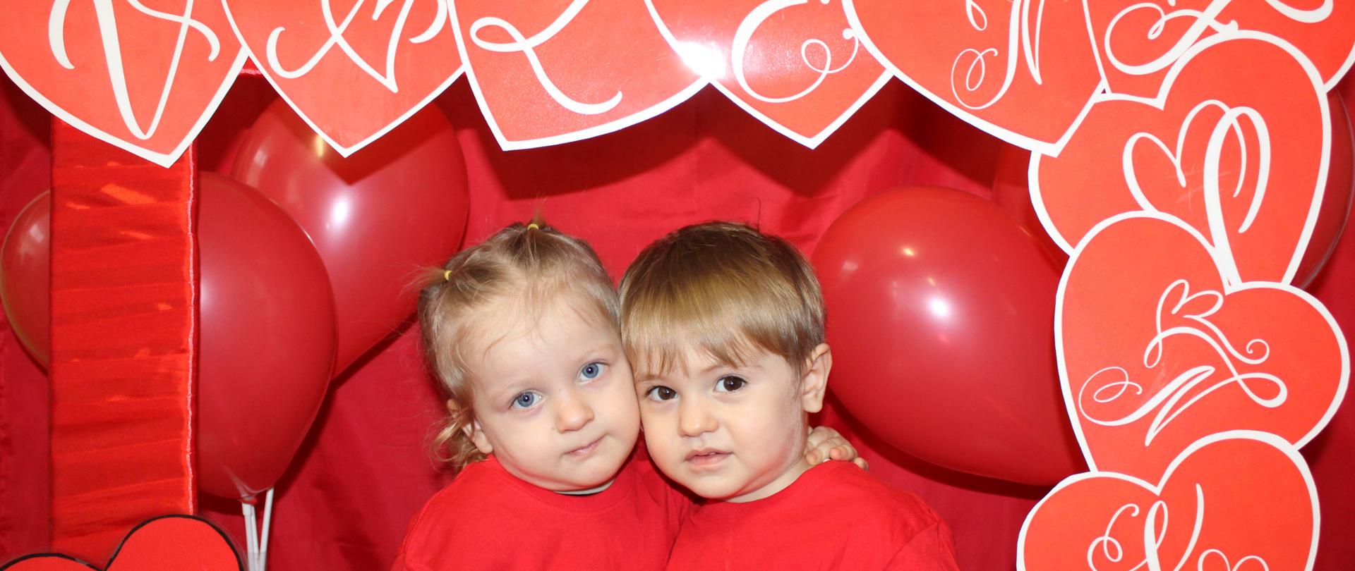 Na zdjęciu widać dójękę dzieci w czerwonej ramce, z napisem Walentynki, w koło rami przyczepione sa czerwone serduszka i wszystko jest na czerwonym tle.