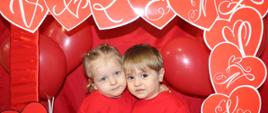 Na zdjęciu widać dójękę dzieci w czerwonej ramce, z napisem Walentynki, w koło rami przyczepione sa czerwone serduszka i wszystko jest na czerwonym tle.