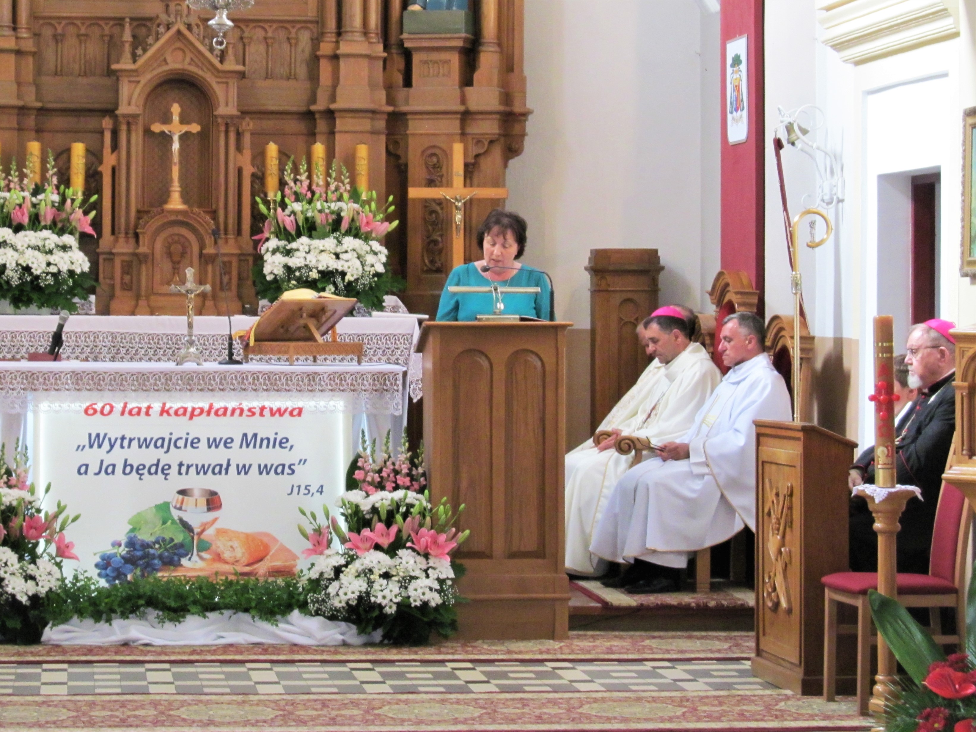 Na zdjęciu moment składania życzeń i gratulacji przez Starostę Elżbietę Sadowską. Widok na drewniany ołtarz kościoła w Niecieczy. Na jego froncie stoją biało - różowe kwitnące kwiaty oraz baner z napisem 60 lat kapłaństwa i cytatem z Pisma Świętego "Wytrwajcie we mnie, a ja będę trwał w Was..." J15, 4. W tle drewniany ozdobny ołtarz. Po prawej stronie widać siedzących księży celebrujących Mszę Świętą, a wśród nich biskupa drohiczyńskiego Piotra Sawczuka oraz biskupa seniora Antoniego Pacyfika - Dydycza.