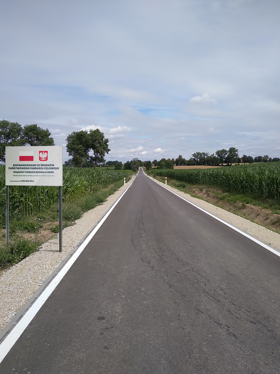 
Droga asfaltowa, po prawej rów, pole i tablica informująca o projekcie, po lewej krzewy i drzewa