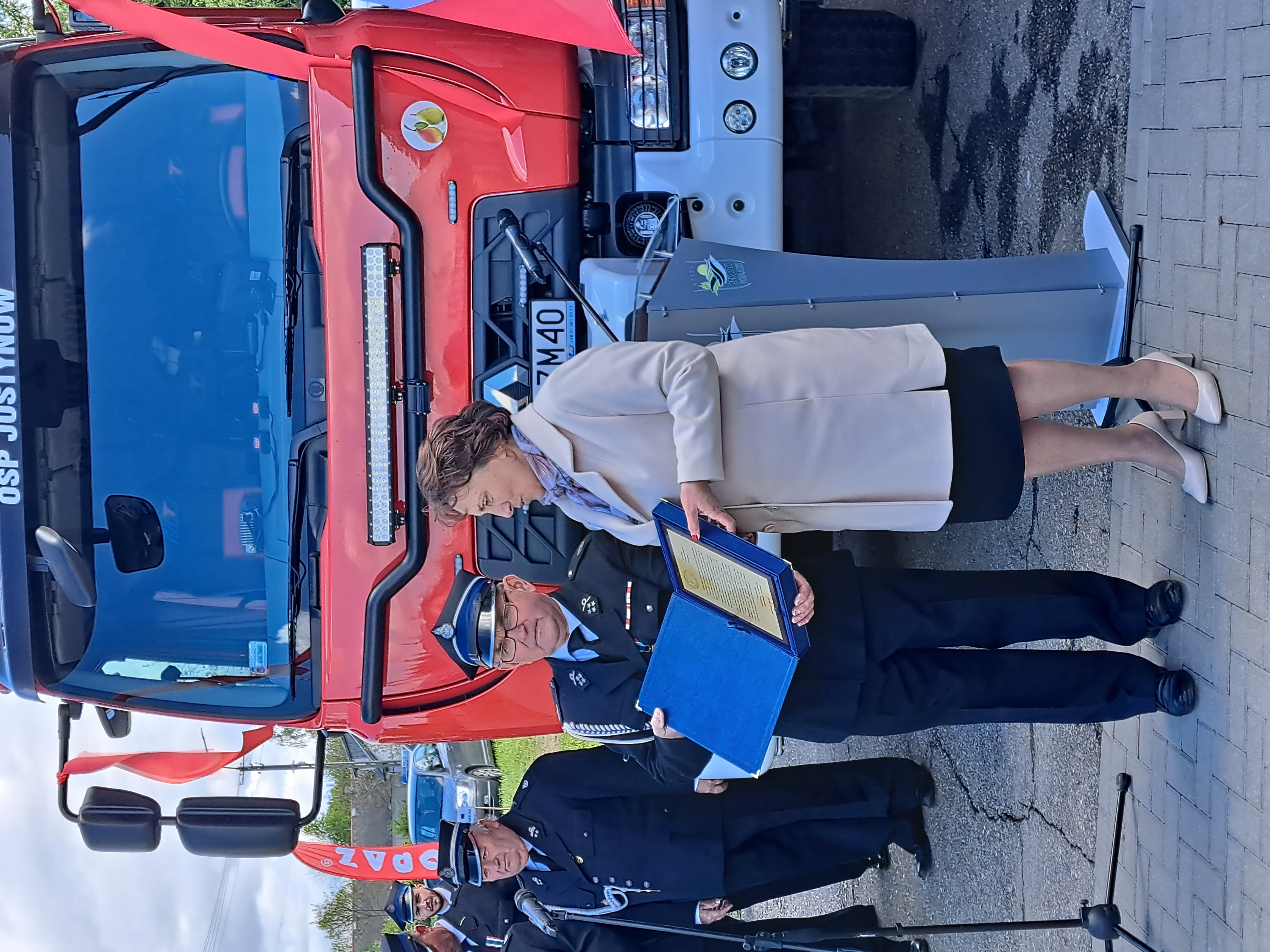 Na zdjęciu starosta Elżbieta Sadowska przekazuje pamiątkowy dyplom na drewnianym podkładzie, z życzeniami dla Druhów strażaków z jednostki OSP w Justynowie. W tle nowy samochód gaśniczo - ratowniczy.