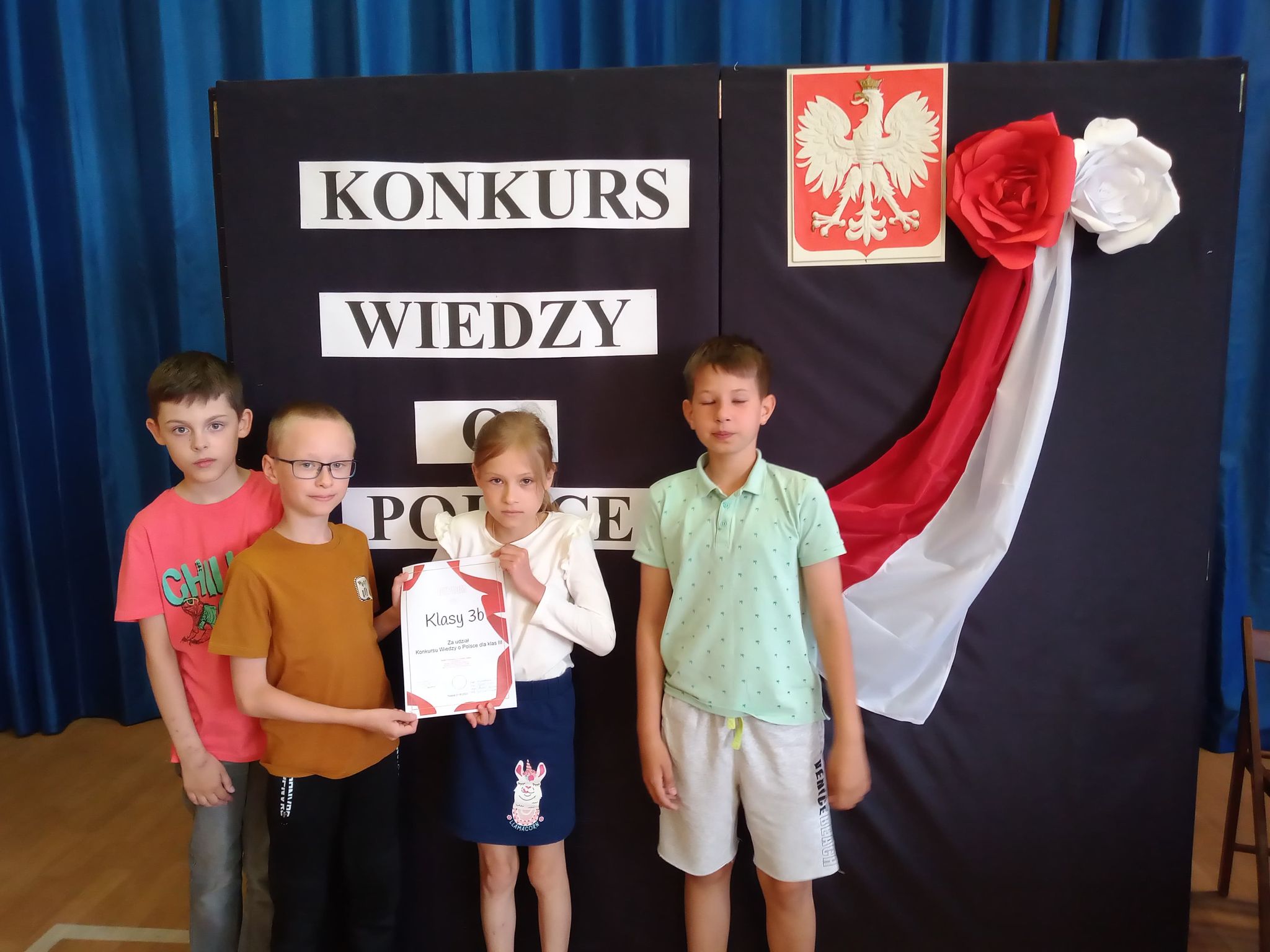 Czworo dzieci. Po środku dziewczynka i chłopiec trzymają w rękach dyplom. Za nimi godło Polski i napis Konkurs wiedzy o Polsce