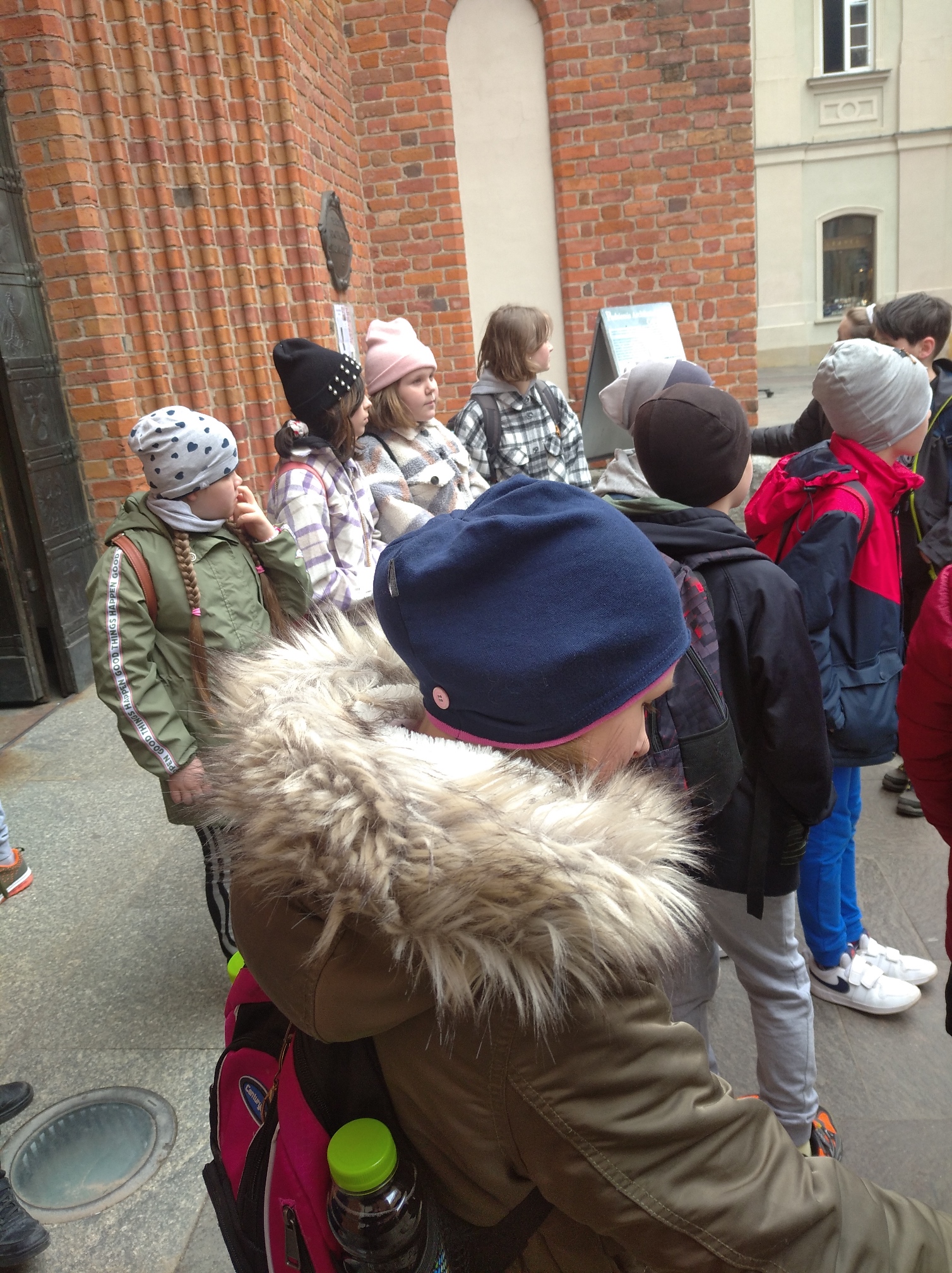 Dzieci ubrane w kurtki, z czapkami stojące przed budynkiem z czerwonej cegły