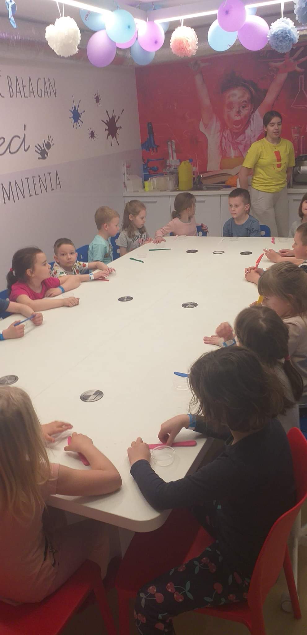 Dzieci siedzą przy długim biały stole. Przed mini leżą plastikowe noże. Obok chodzi kobieta w żółtej bluzce