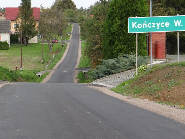 Przebiegająca przez lekko pofałdowany krajobraz droga o nawierzchni asfaltowej. Na pierwszym planie, na poboczu, po prawej stronie tablica z nazwą miejscowości Kończyce Wielkie. 