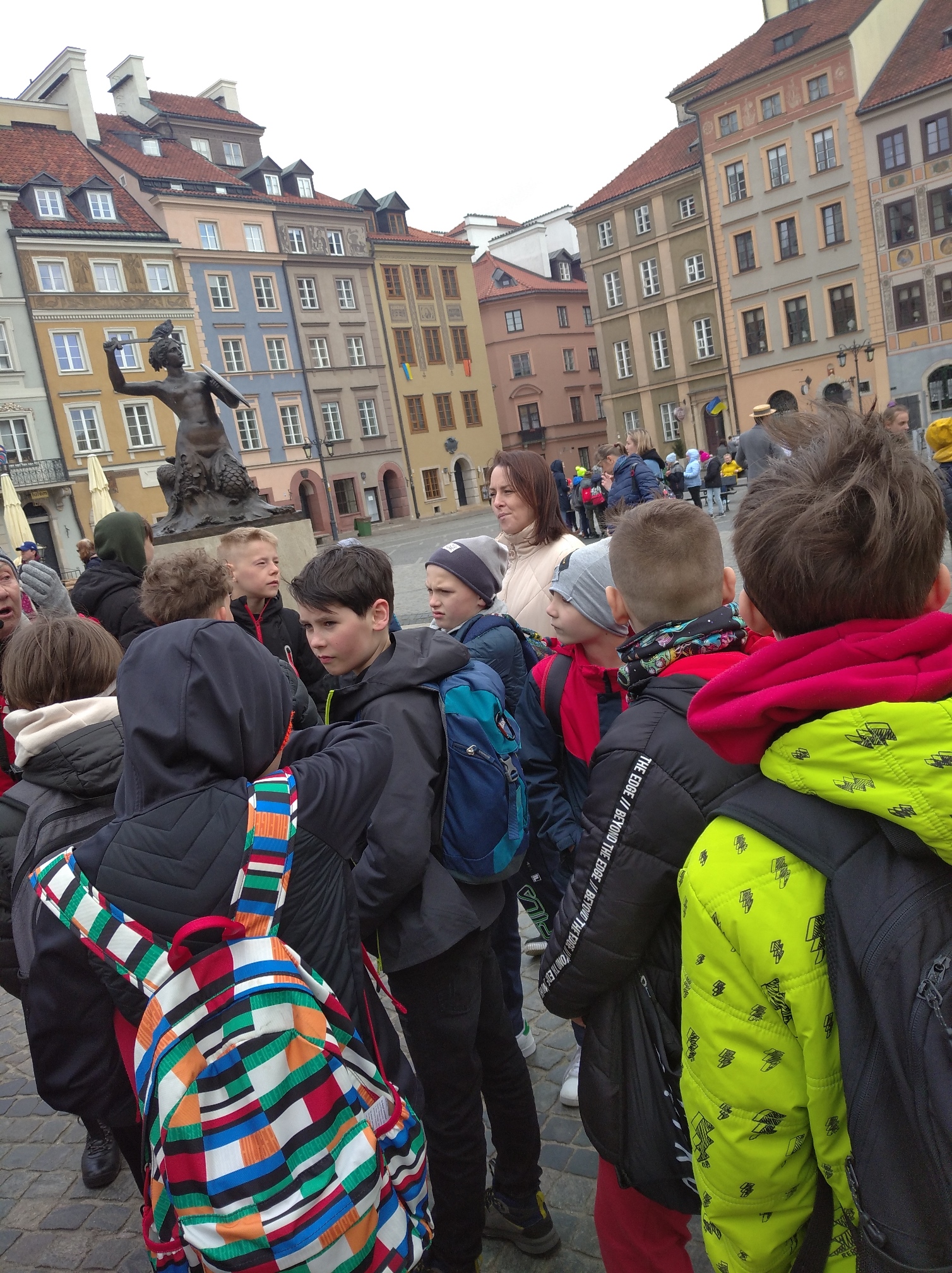 Dzieci na placu na którym stoi posąg pół kobiety pół ryby z mieczem w ręku. Wokół stoją kolorowe budynki