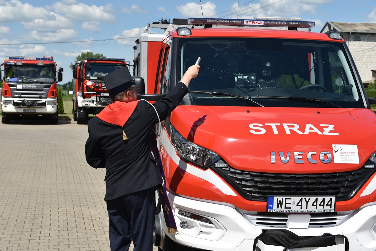 Ksiądz kapelan strażaków Krzysztof Kisielewicz kropi wodą święconą nowy lekki samochód ratownictwa technicznego. W tle ustawione przodem ciężkie samochody strażackie w kolorze czerwonym.