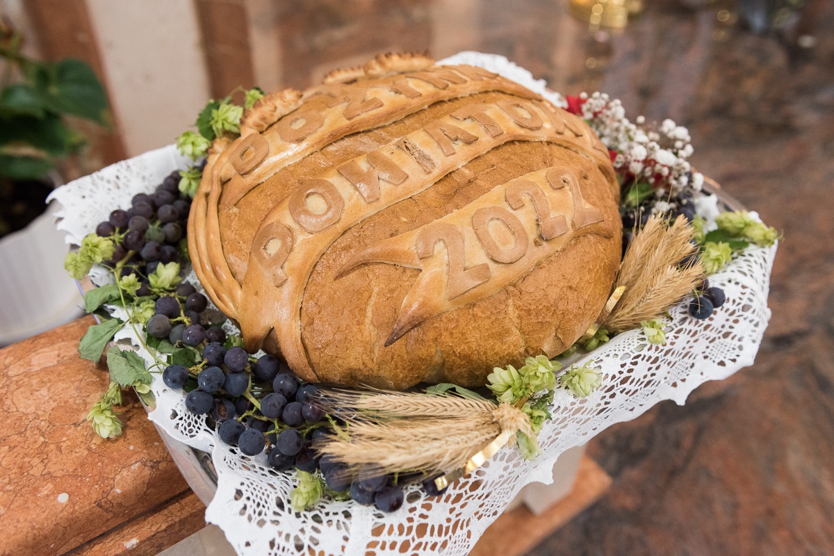 Na zdjęciu widać okrągły pszenny chleb z napisem dożynki powiatowe 2022 przyozdobiony winogronem, kłosami zbóż, chmielem oraz kwiatami w kolorze białym i czerwonym