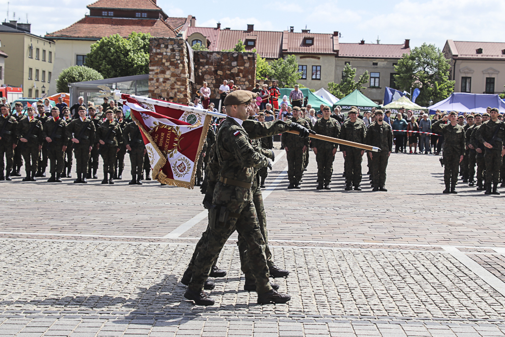 zdjęcie przedstawia w tle stojących w szeregach żołnierzy. Przed nimi troje żołnierzy niesie flagę ze sztandarem.