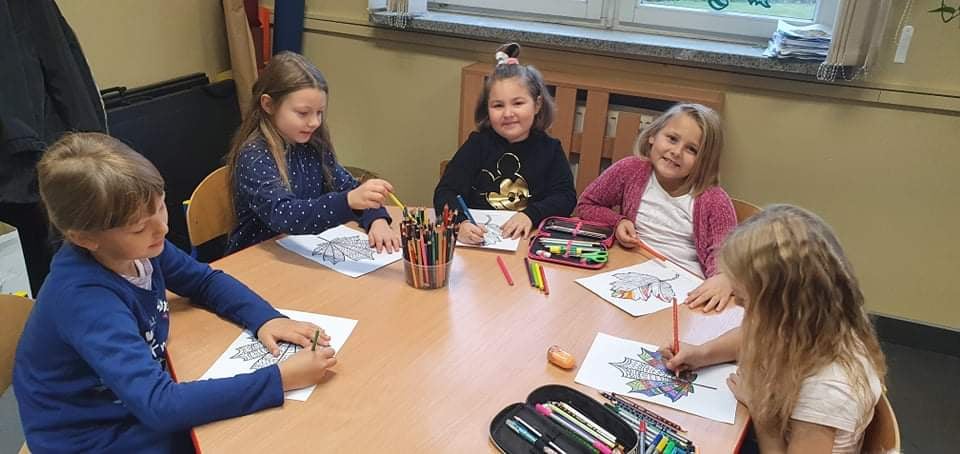 Dzieci siedzą przy stoliku, na którym leżą piórniki i kredki. Dzieci kolorują obraki liści