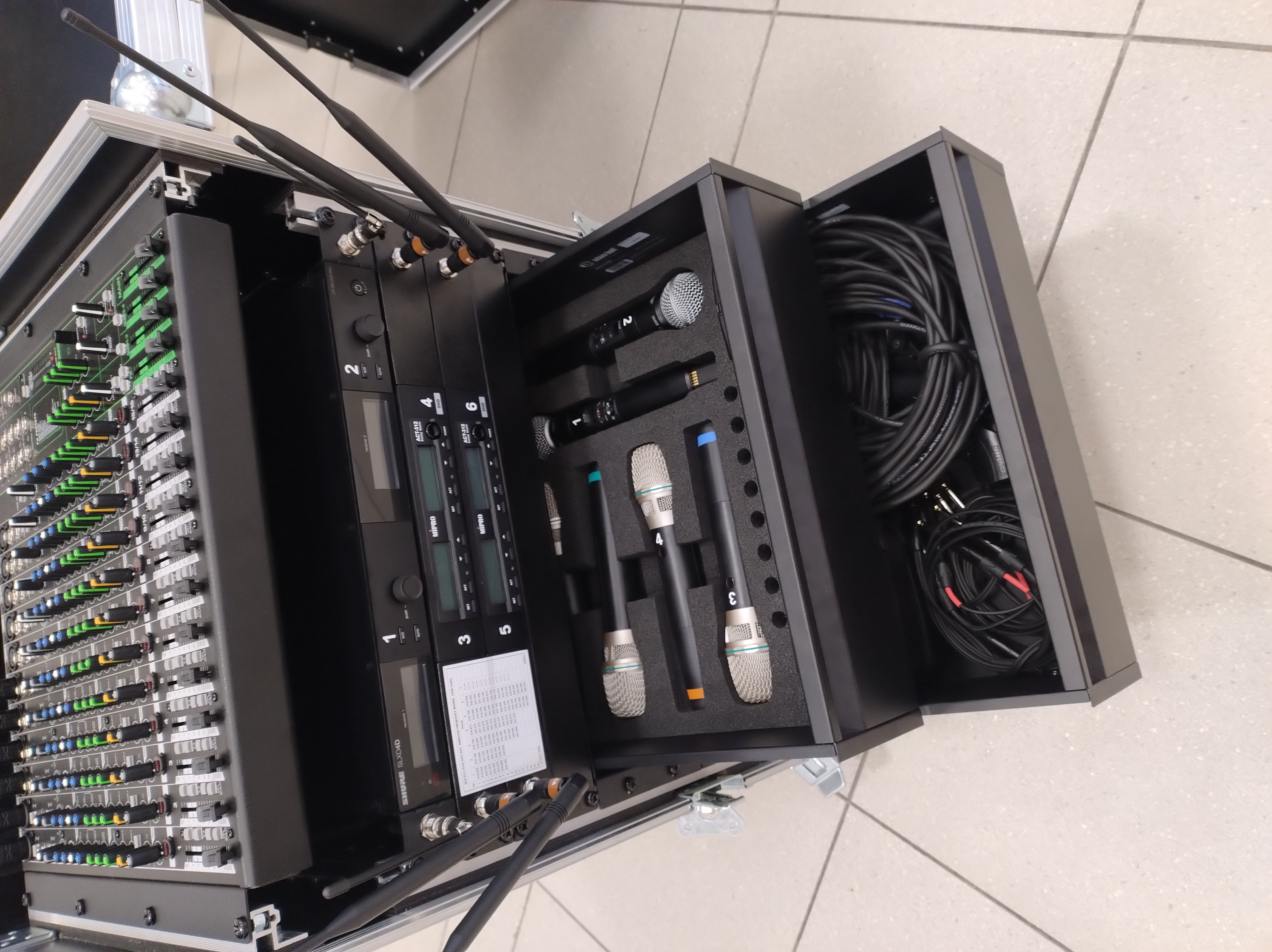 Zdjęcie przedstawia profesjonalny system nagłośnienia i mikrofonów, mikser audio, odbiorniki, mikrofony ręczne oraz akcesoria. Sprzęt znajduje się w mobilnej szafie transportowej.