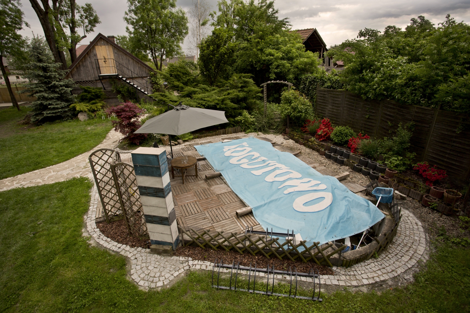 Basen zakryty niebieską folią z napisem Kogutowo w tle trawnik, drzewa i ściana drewnianego domu ze schodami