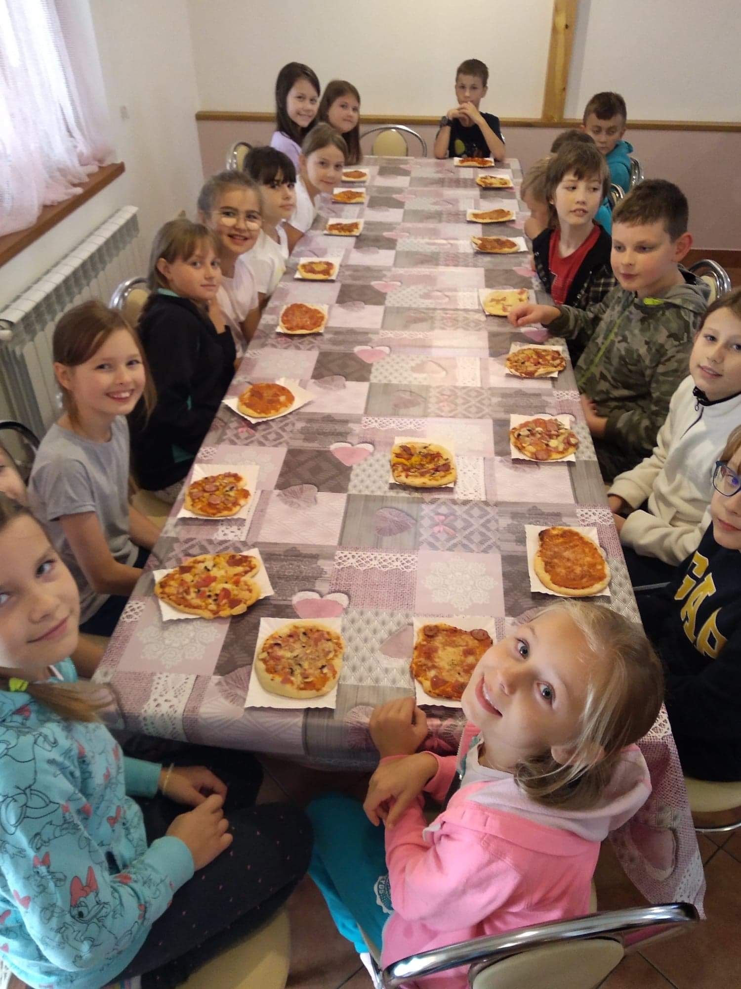 Dzieci siedzą przy długim stole. Przed każdym z nich leży pizza