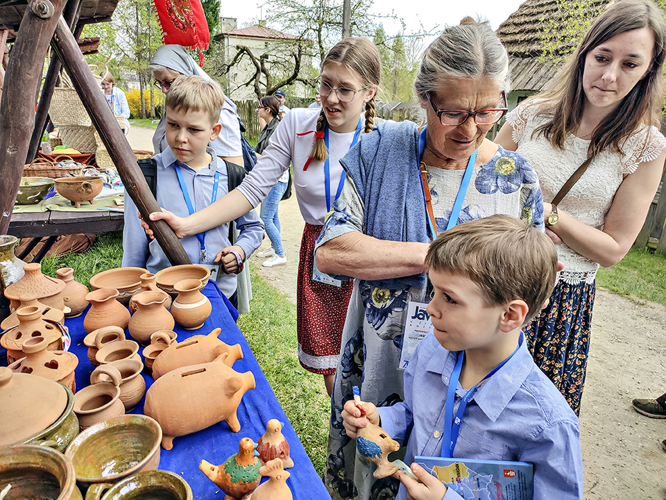 Zdjęcie przedstawia grupę osób (dzieci i wraz z babcią) odwiedzających stoisko z wyrobami ceramicznymi. 