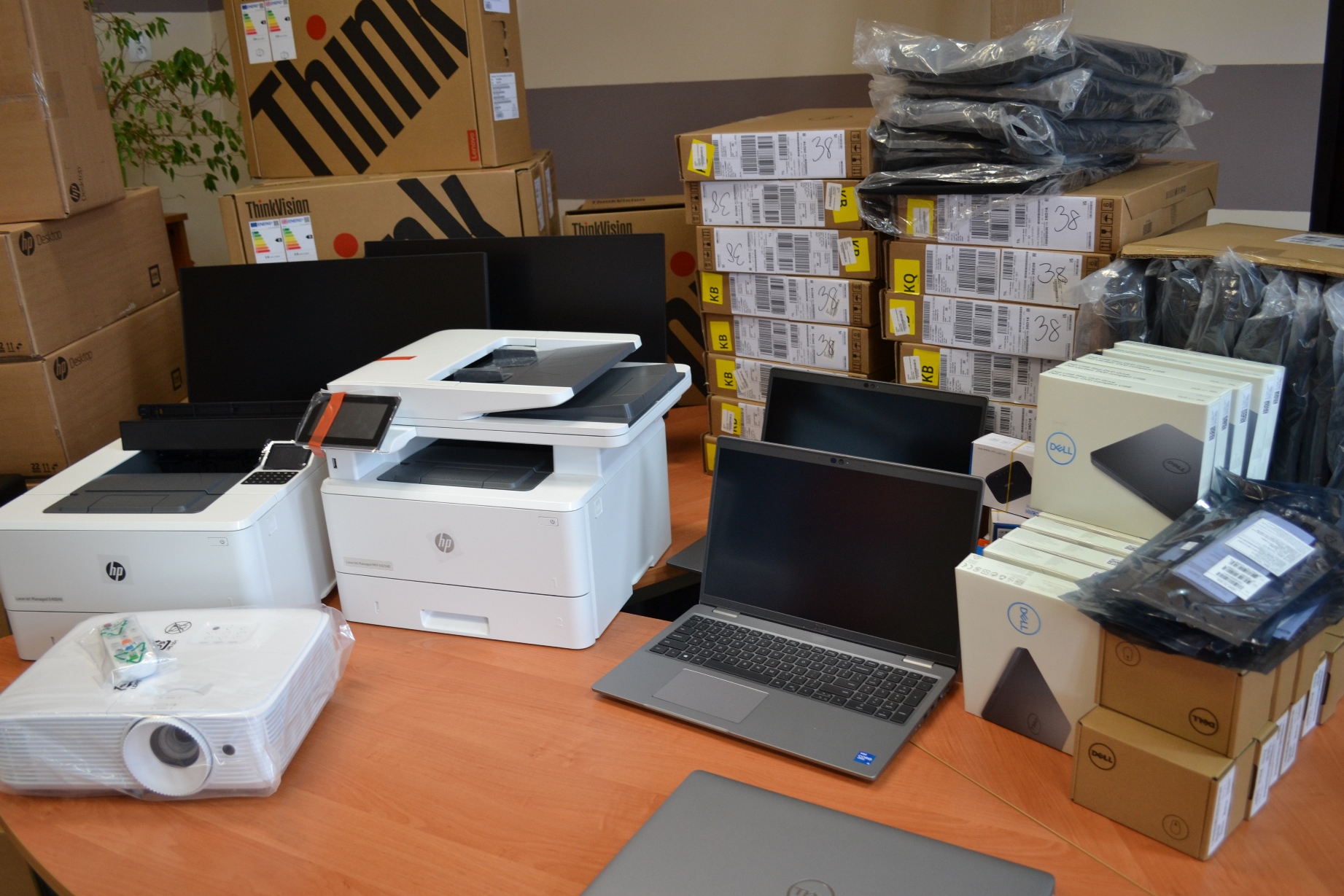 Na stole leżą drukarki, laptopy, projektor oraz kartony