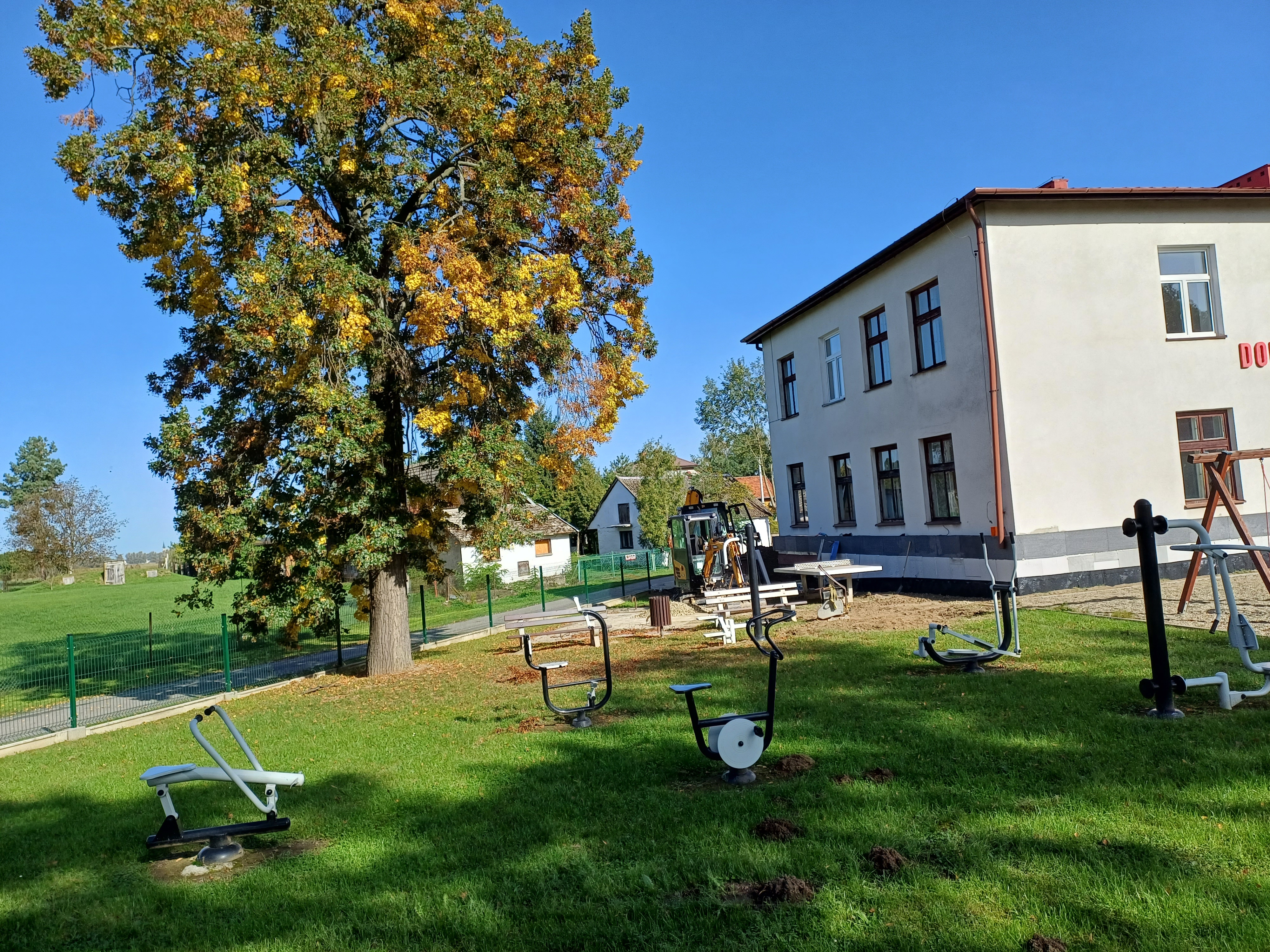 Zdjęcie przedstawia fragment budynku, z lewej strony rośnie drzewo. Przed budynkiem znajdują się sprzęty do ćwiczeń plenerowych.