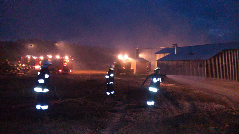 Troje strażaków w strojach z pasami odblaskowymi w nocy. Strażak po prawej trzyma wąż z którego leci woda. W tle samochody strażackie z włączonymi oświetleniem terenu