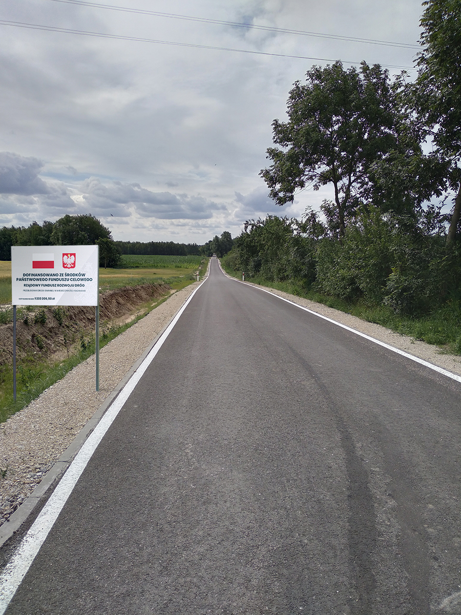 Droga asfaltowa, po prawej rów, pole i tablica informująca o projekcie, po lewej krzewy i drzewa