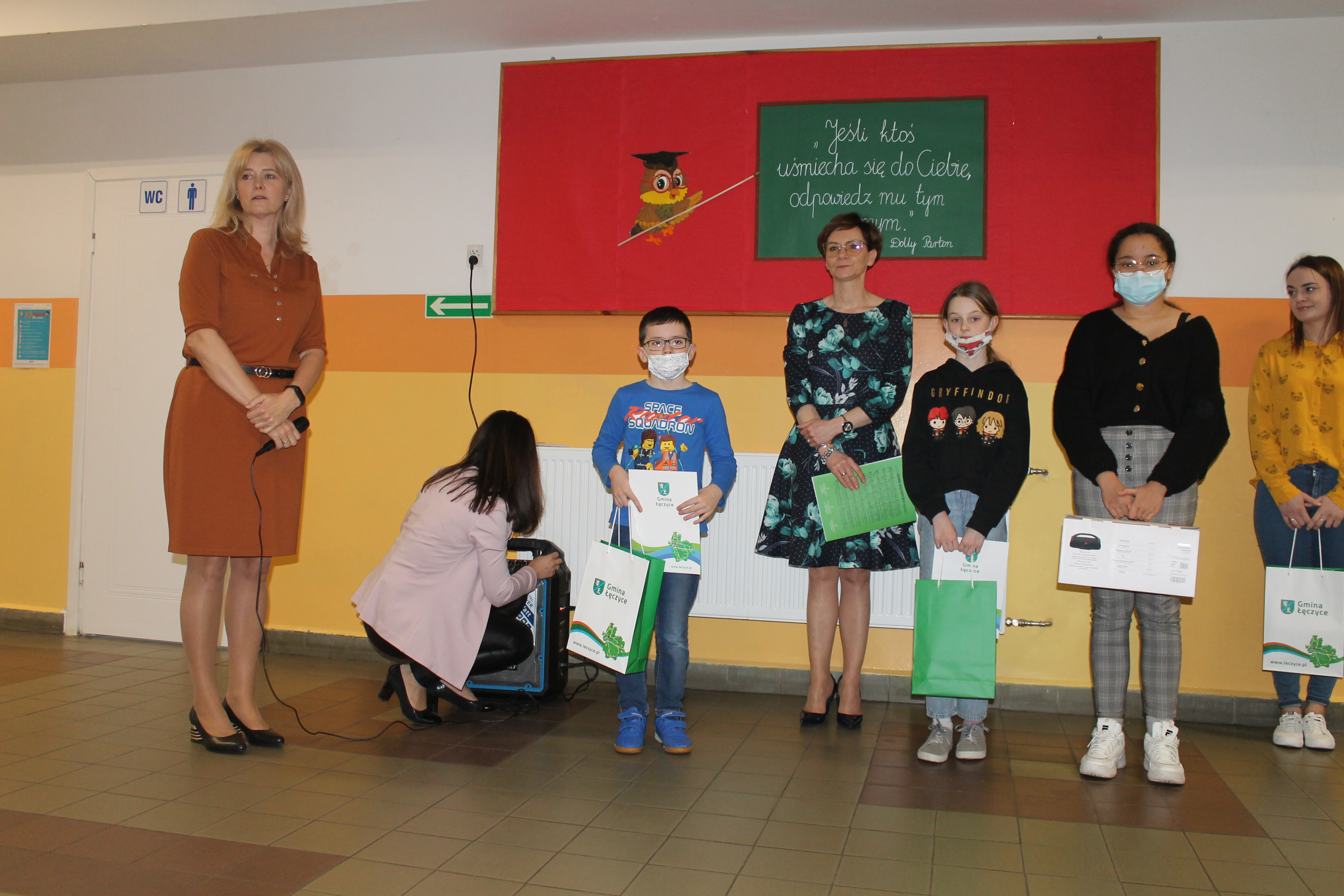 Zdjęcie przedstawiające dzieci oraz dorosłych na wręczeniu nagród w konkursie, w tle wisząca ilustracja sowy wskazującej na tekst: "Jeśli ktoś uśmiecha się do Ciebie, odpowiedź mu tym samym"