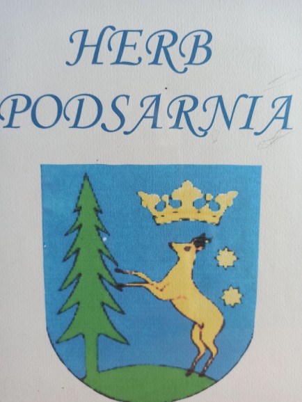 Herb Podsarnia przedstawiający na niebieskim tle zielony smrek, przy którym na dwóch nogach stoi sarna.