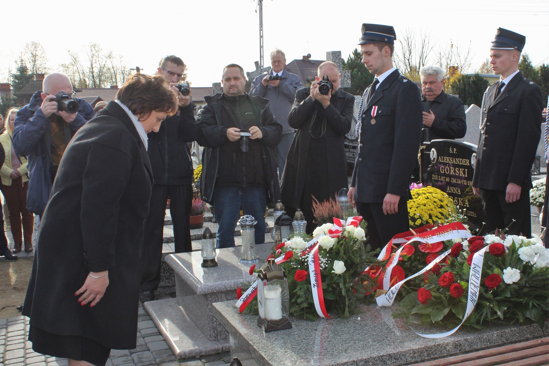 Moment złożenia przez starostę kwiatów na pomniku ks. Antoniego Łapaya. Starosta kłania się stojąc na baczność przed miejscem pochówku księdza. W tle fotoreporterzy oraz uczestnicy wydarzenia.