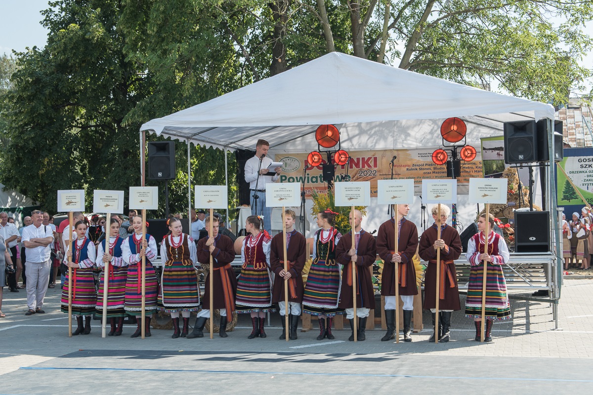 Na zdjęciu widać scenę dożynkową, a przed nią ustawieni w strojach ludowych członkowie Zespołu Pieśni i Tańca Sokołowianie trzymający tabliczki z nazwami poszczególnych delegacji wieńcowych.
