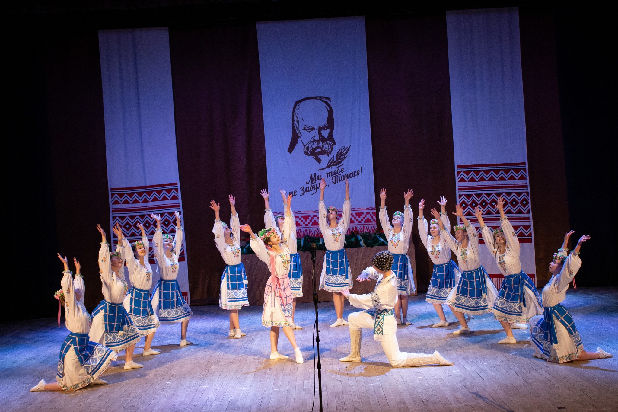 zdjęcie przedstawia zespół taneczny składający się z jednego mężczyzny oraz kilkunastu kobiet ubranych w stroje regionalne w trakcie występu na scenie