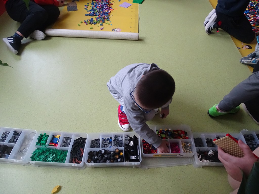 Zdjęcie przedstawia małego uczestnika zimowych półkolonii, który dobiera klocki z pudełka w celu upiększenia swojego domku z klocków LEGO. 