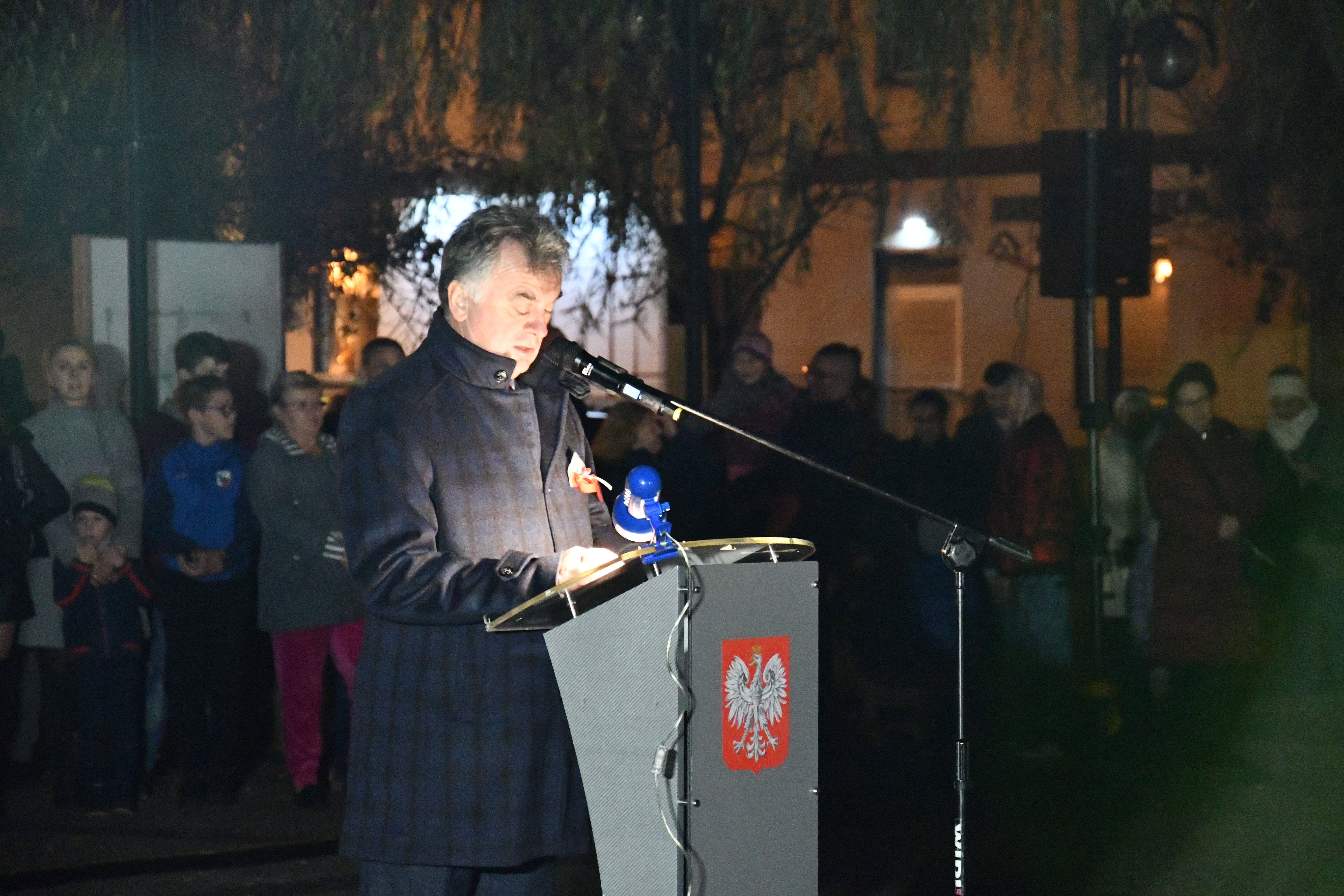 Burmistrz Janusz Piechocki podczas przemówienia, stoi przy mównicy, za nim zebrał się tłum słuchaczy i uczestników wydarzenia.