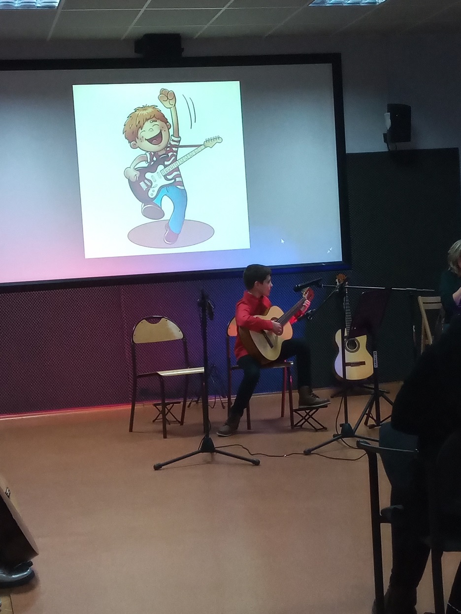 Chłopiec siedzi na krześle z gitarą w rękach, za nim wyświetla się obrazek chłopca z gitarą